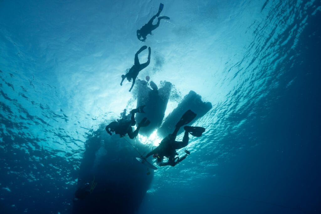 Zanzibar Snorkeling and Safari Packages | Dive into Tanzania’s Best 3 Silhouetted divers swimming beneath a yacht in clear ocean waters.