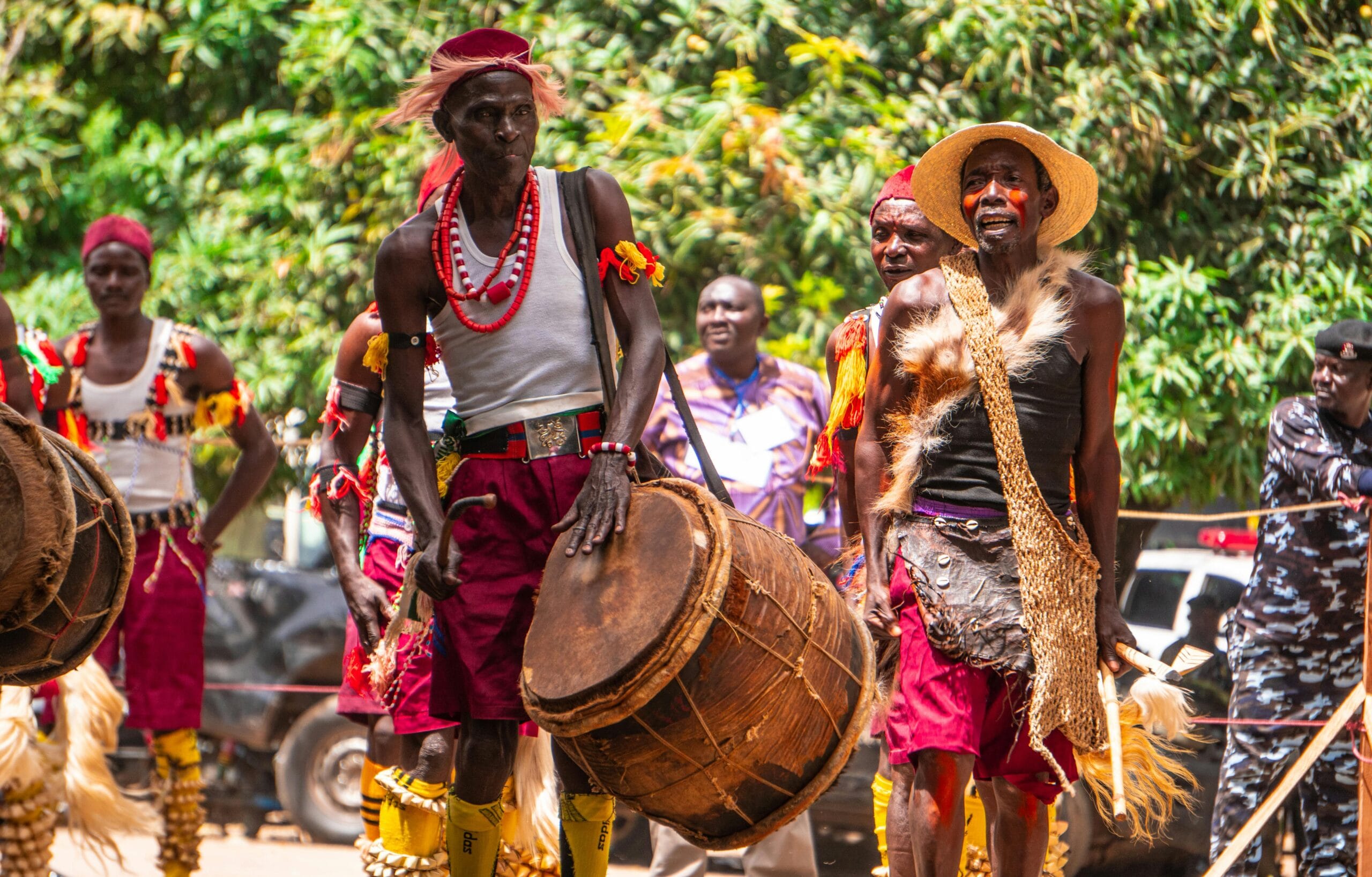 Group of African men in traditional attire performing with drums during a vibrant outdoor cultural festival.