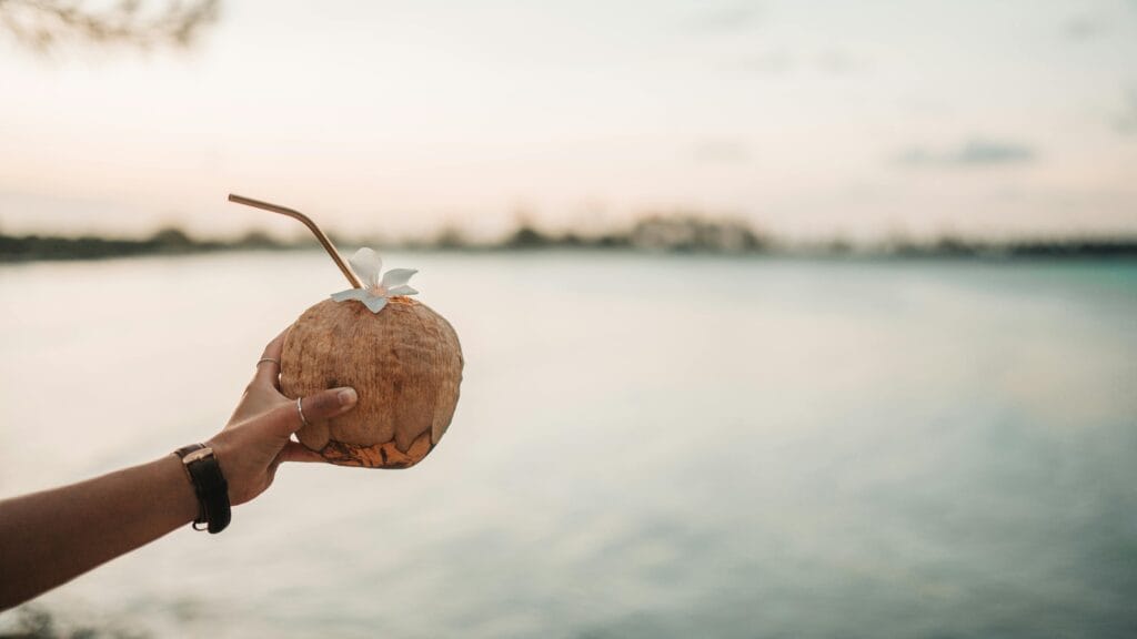 Hand holding a tropical coconut drink with a straw on Zanzibar's serene coastline.