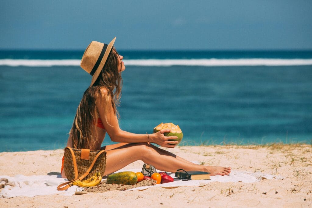 A woman in a swimsuit and sun hat relaxes on a sandy beach, holding a coconut, with the ocean in the background.