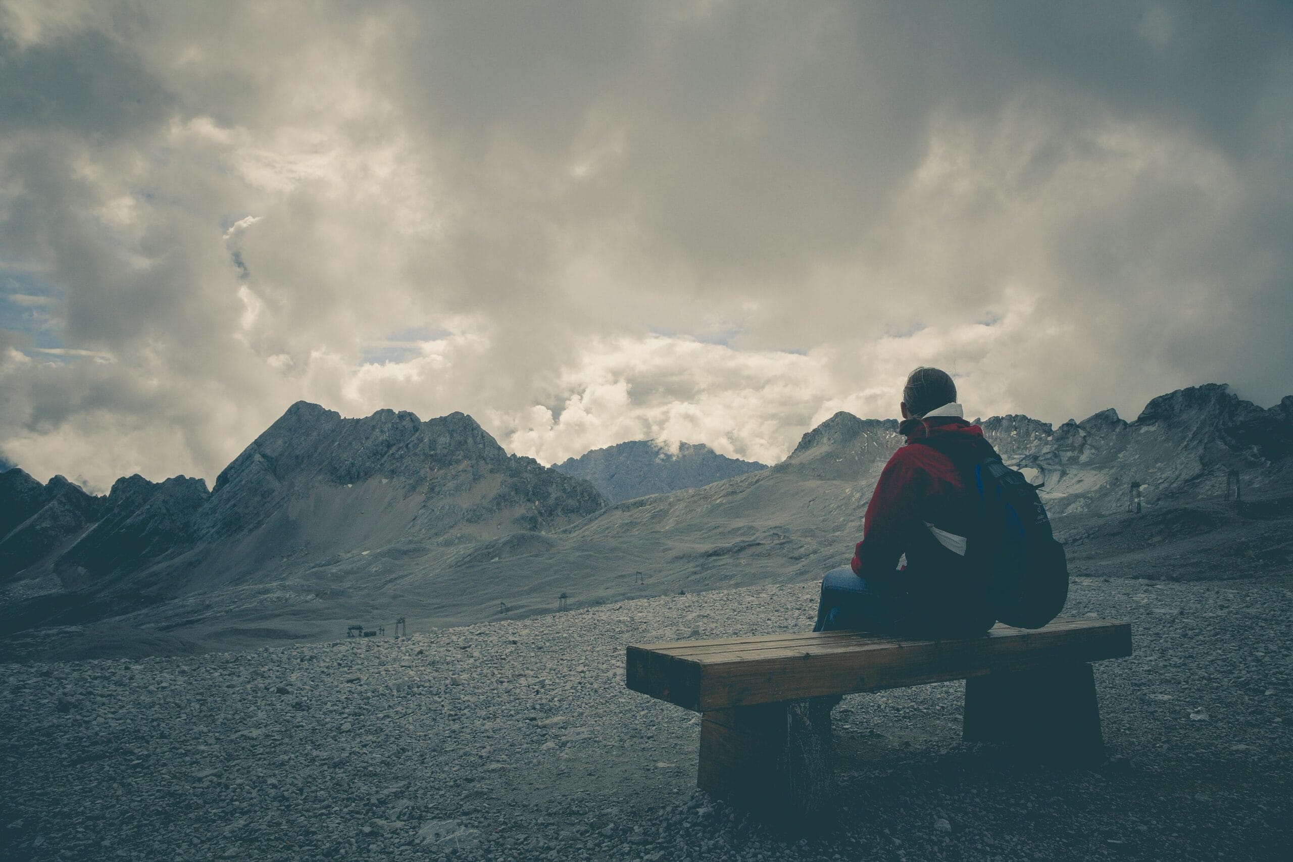 A solitary traveler sits on a bench, gazing at a mountain range under a cloudy sky.