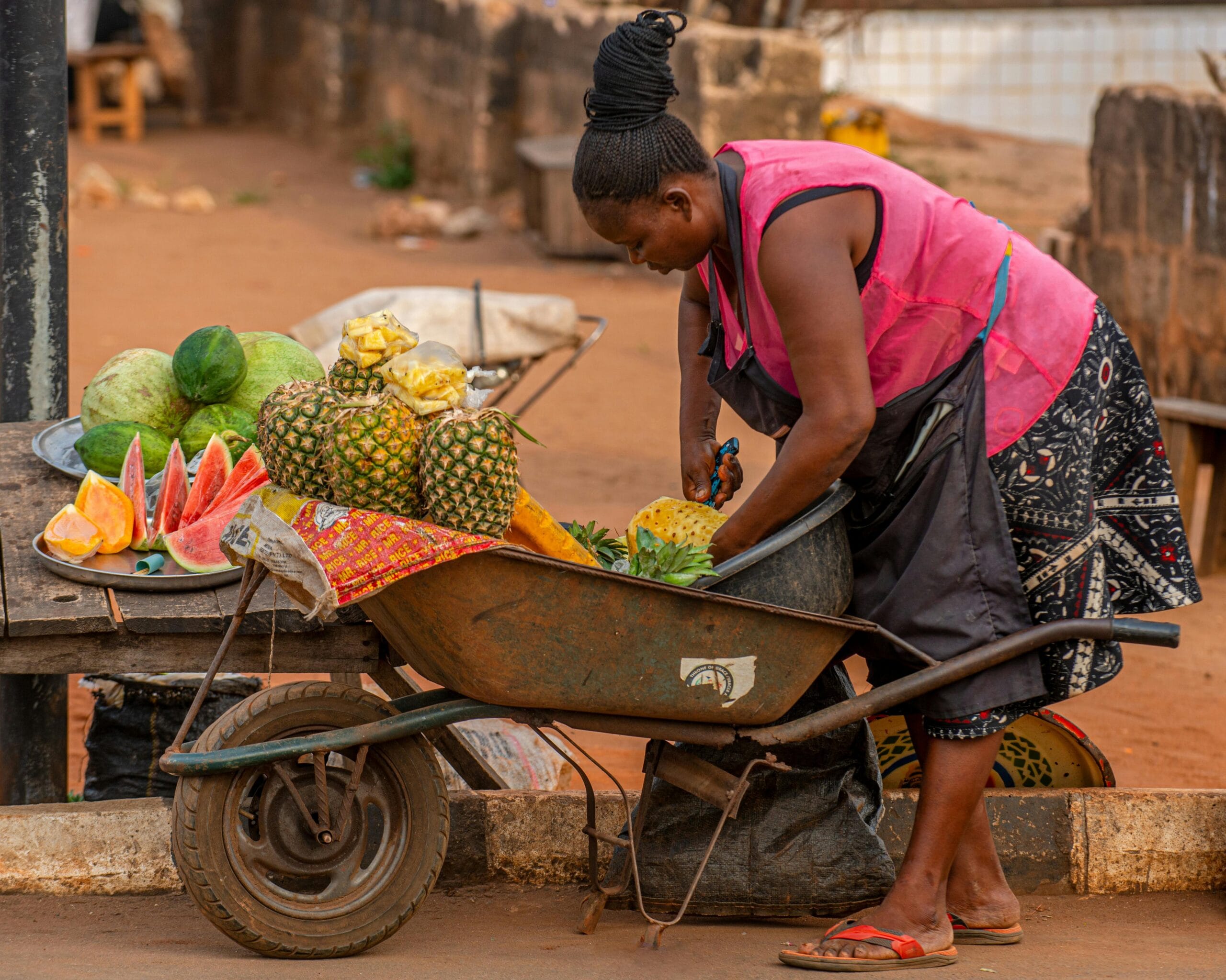 African woman preparing fresh pineapples on a street vendor wheelbarrow in a local market.