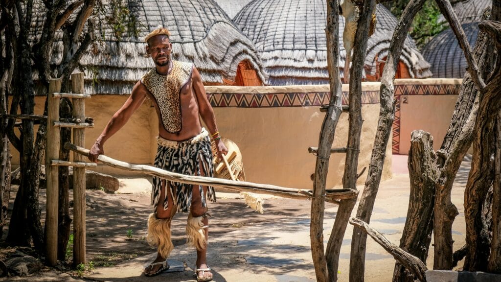 A man in traditional attire poses outdoors in a South African tribal setting.