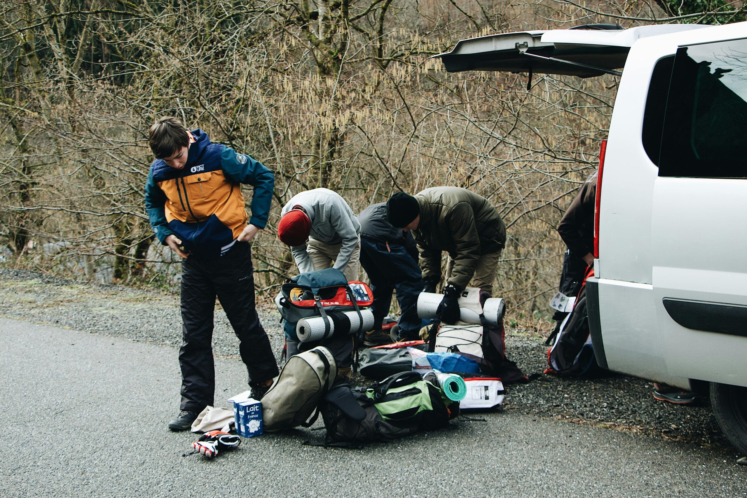 A group of travelers prepares their gear for an outdoor hiking adventure by their vehicle on a scenic road.