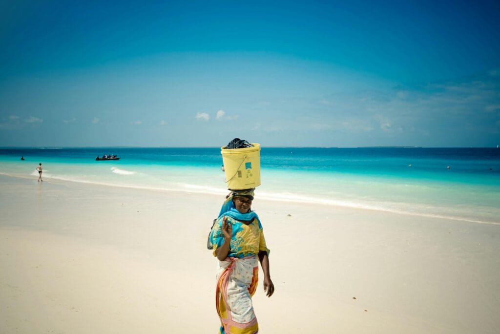 An African woman carries a bucket on a pristine Zanzibar beach under a clear blue sky.