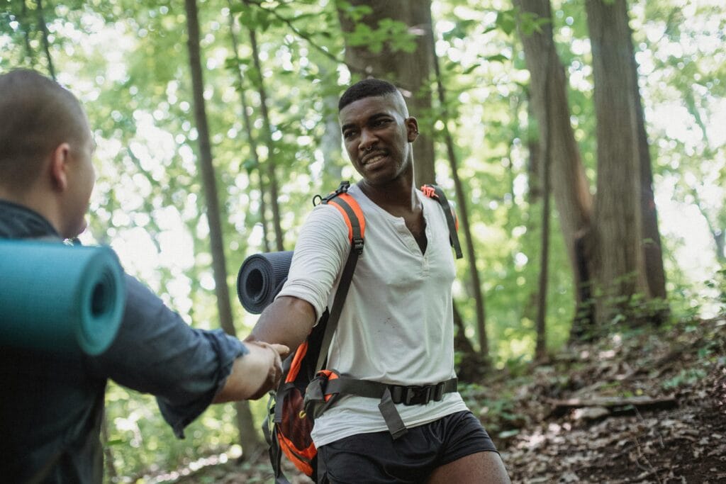 Two men hiking in a lush forest, one reaching out to help the other on a path.
