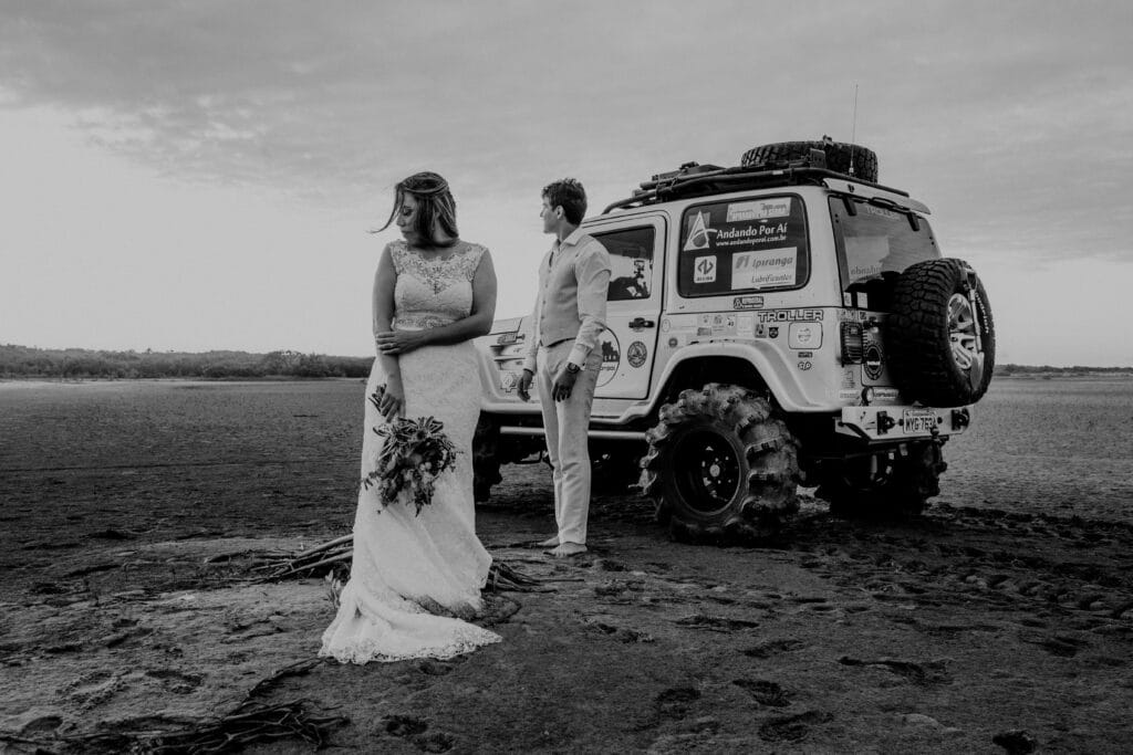 Newlywed couple in elegant attire with SUV in desert, embracing love and exploration.