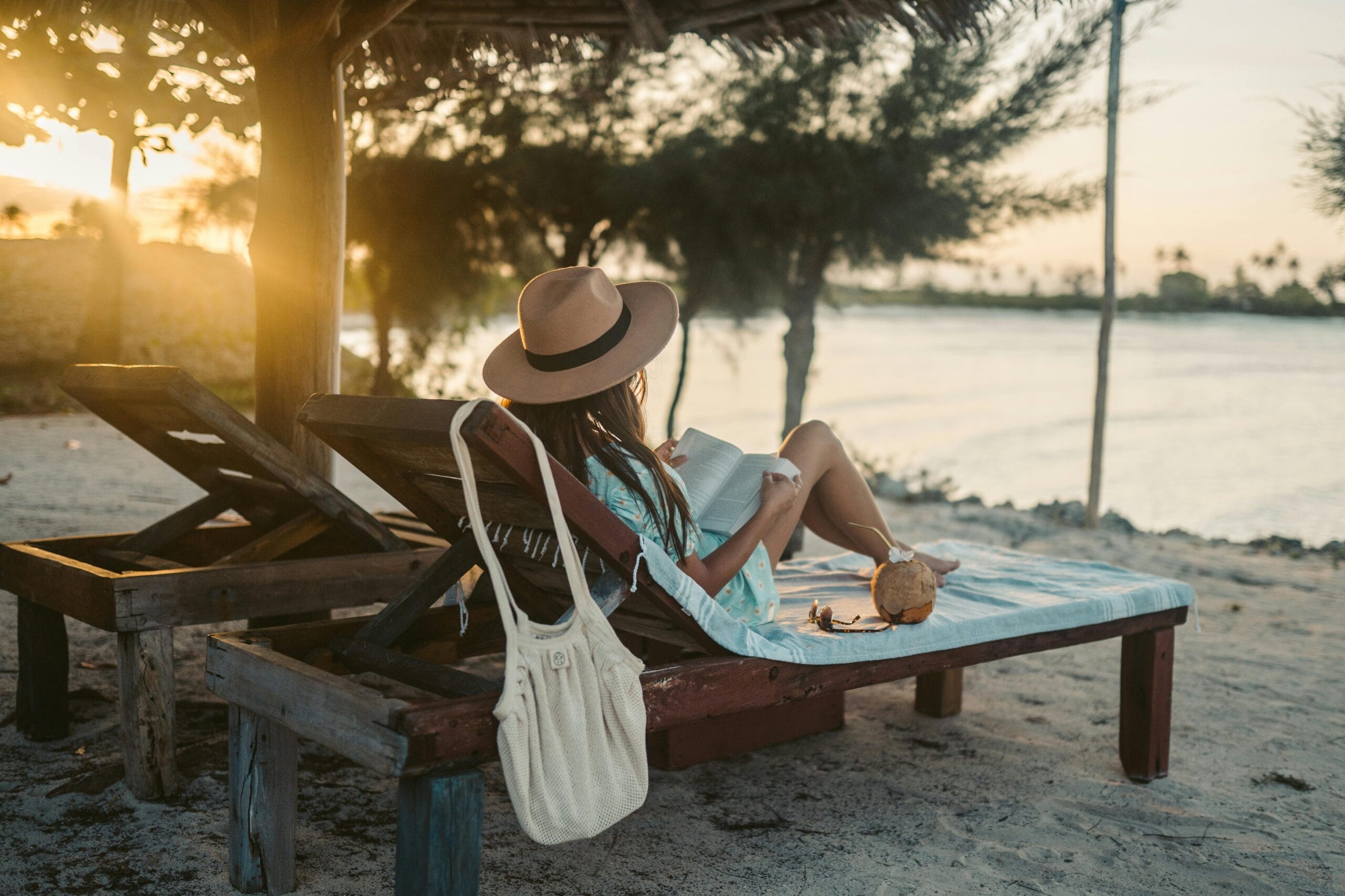 Woman reading on a beach lounger under a sun hat in Zanzibar at sunset, embracing vacation tranquility.