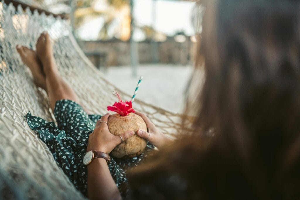 A woman enjoys a coconut drink while relaxing in a hammock on a sandy beach.