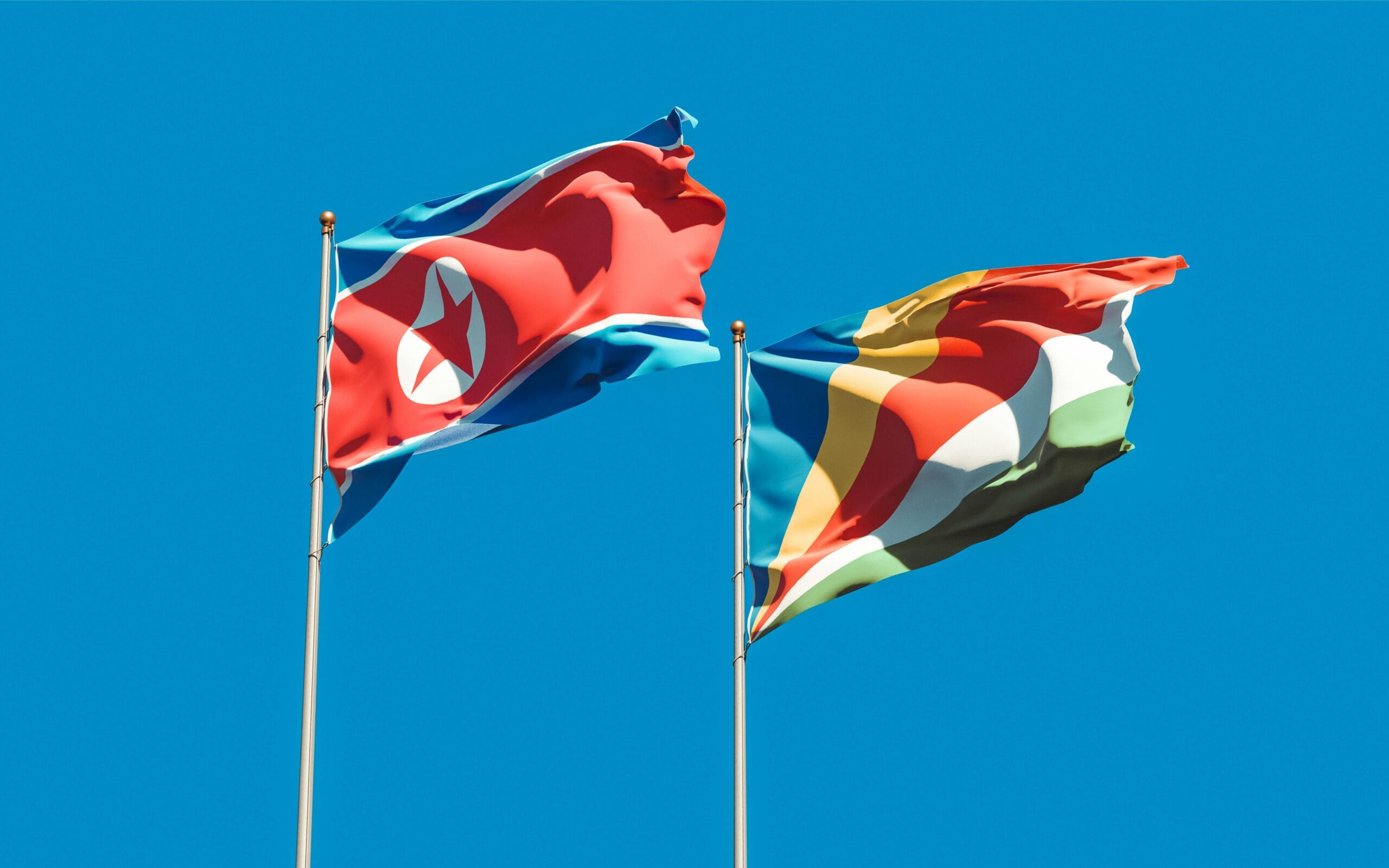 Vibrant national flags of North Korea and Seychelles on flagpoles waving under a clear blue sky.