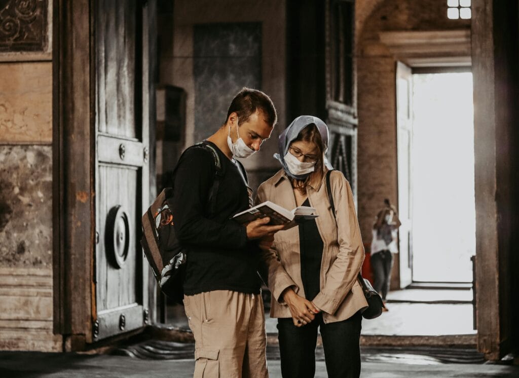 A couple wearing face masks reads a book together in a historic building.