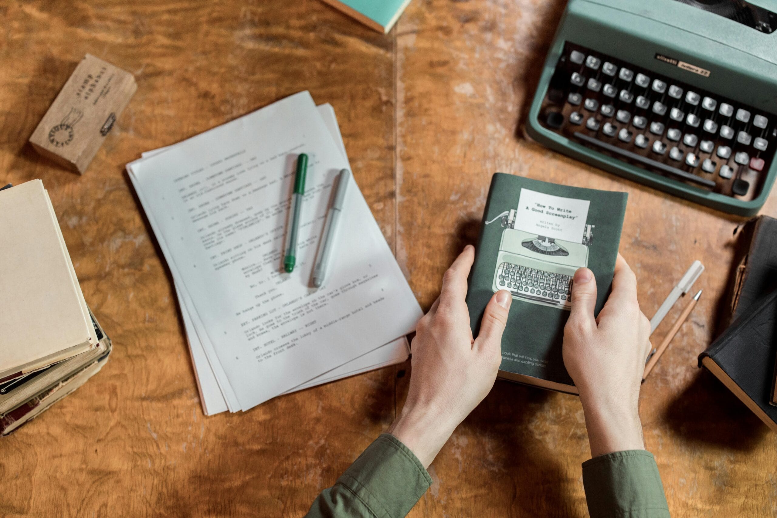 A vintage setup showcasing a typewriter, manuscripts, and hands holding a book on writing.