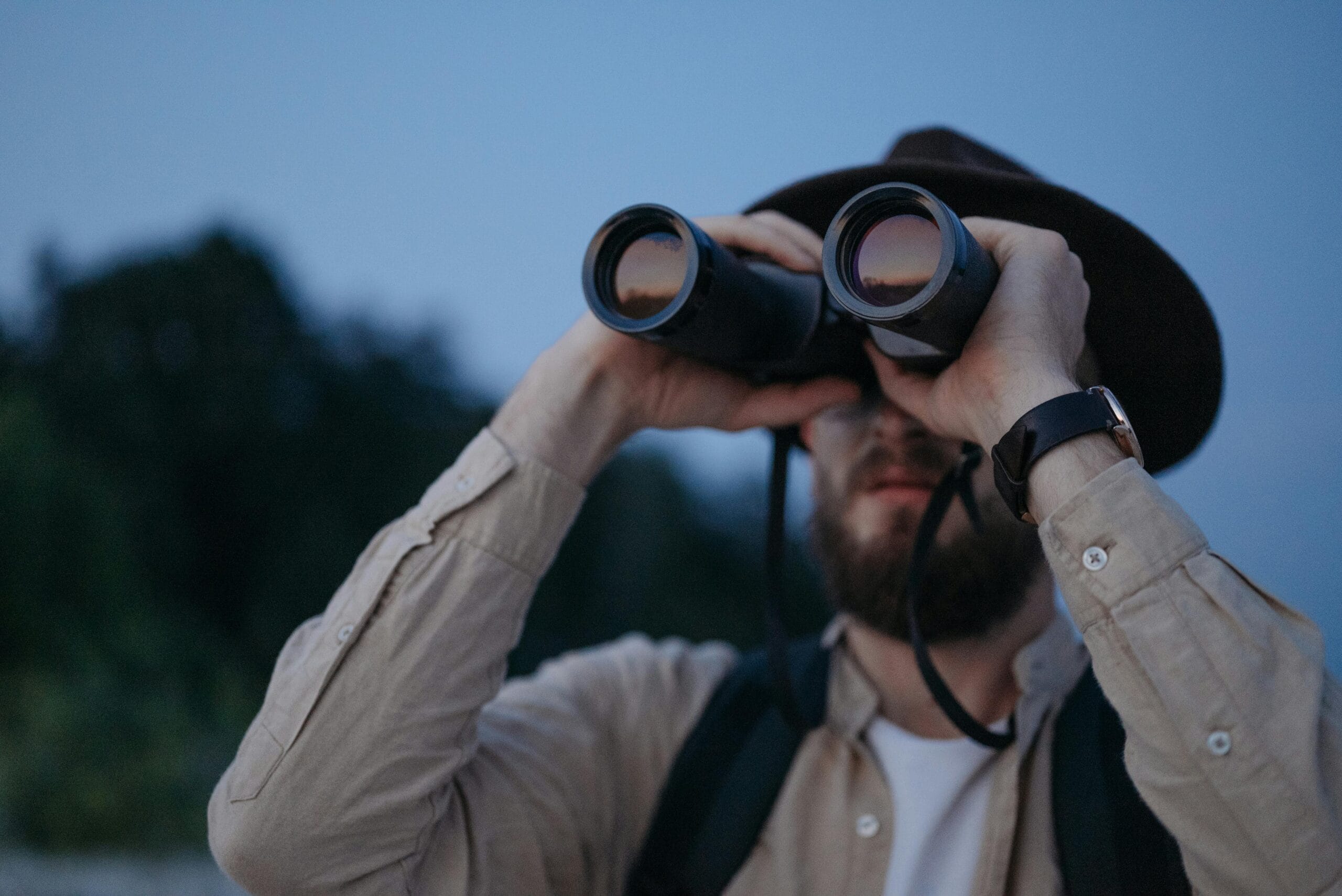 A man wearing a hat uses binoculars to observe the outdoors during twilight in a natural setting.