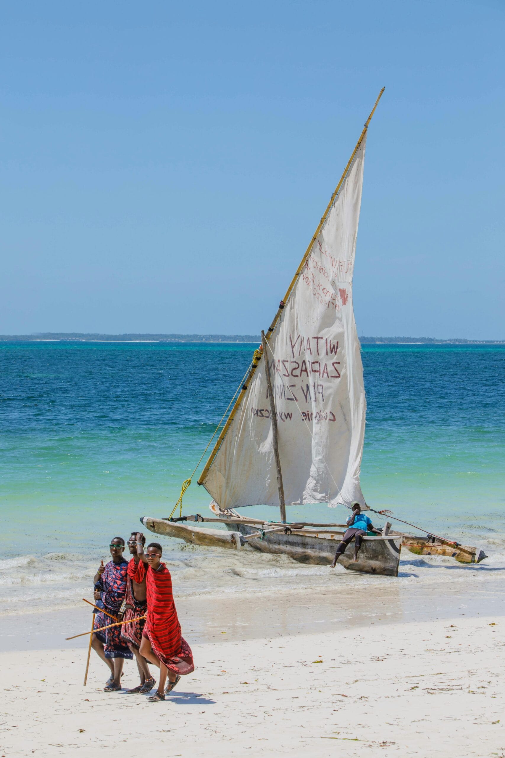 A group of Maasai people walking by a traditional sailboat on a sunny tropical beach.